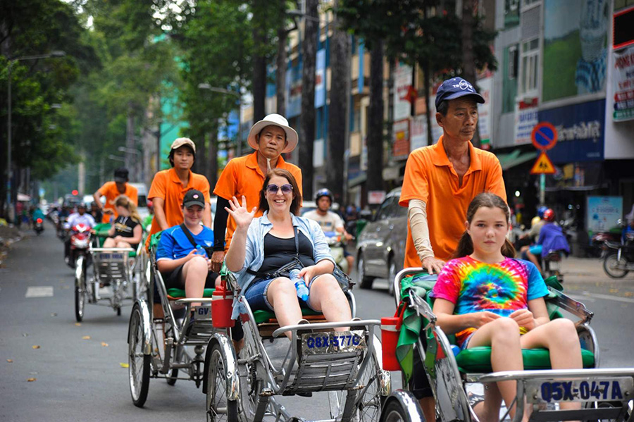 Experience Ho Chi Minh City’s vibrant streets up close! A classic trishaw ride offers a unique perspective of Saigon’s bustling daily life. People enjoying a trishaw ride through a busy street in Ho Chi Minh City, Vietnam, with local buildings visible.